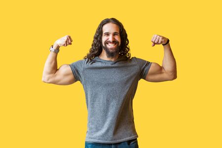 Portrait of happy strong bearded young bodybuilder man with long curly hair in grey tshirt standing, posing and looking at camera with toothy smile. indoor studio shot isolated on yellow background.の写真素材