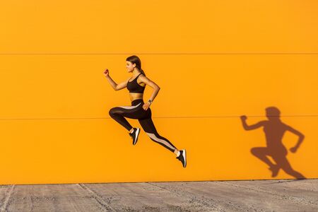 Young satisfied sporty beautiful woman with fit body jumping and running, hurry up against orange wall background. Gymnast jumping high with toothy smile, full length, outdoorの写真素材