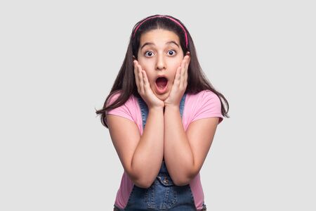 Portrait of surprised beautiful brunette young girl in casual pink t-shirt and blue overalls standing with hands on face and looking with amazed face. studio shot, isolated on light gray background.の写真素材
