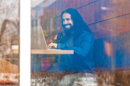 Portrait through the window of handsome young adult man freelancer in casual style sitting in cafe, chating with his friend in laptop, bussinessman in office, toothy smile. outdoor, lifestyle conceptの写真素材
