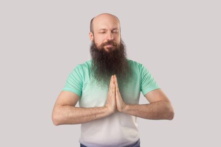 Portrait of calm serious middle aged bald man with long beard in light green t-shirt standing with closed eyes, palm hand and doing yoga meditation. indoor studio shot, isolated on grey background.の写真素材