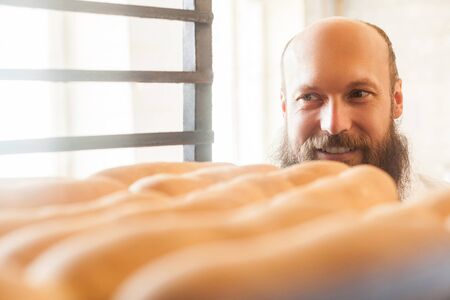 Portrait of satisfied young adult chef baker with long beard in white uniform standing in his bakery...