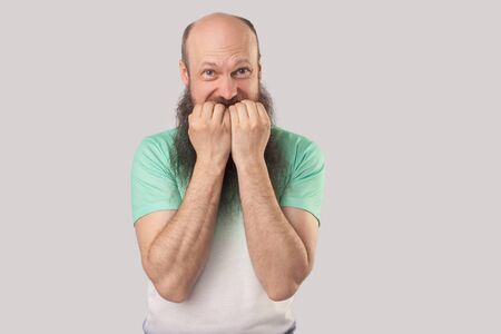 Portrait of nervous middle aged bald man with long beard in light green t-shirt standing, bitting his nails and looking at camera with worry panic face. indoor studio shot isolated on grey backgroundの写真素材