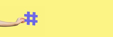 Social media concept. closeup of human hand holding and showing large big blue hashtag sign. indoor studio shot, isolated on yellow background copyspace.の写真素材