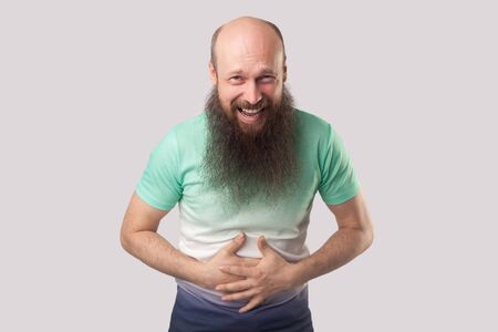 Portrait of funny middle aged bald man with long beard in light green t-shirt standing, holding his belly, laughing, enjoying and looking at camera. indoor studio shot, isolated on grey background.の写真素材