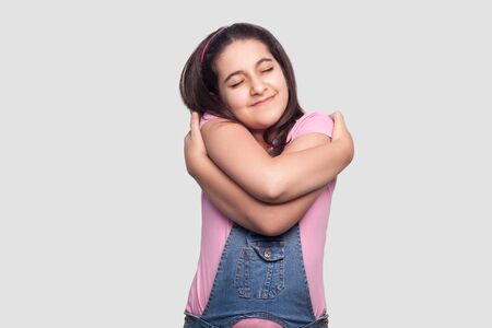 Portrait of happy satisfied beautiful brunette young girl in casual pink t-shirt and blue denim overalls standing, hugging herself and enjoying. indoor studio shot, isolated on light gray background.の写真素材