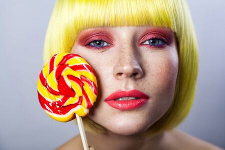 Beauty portrait of cute young female model with freckles, red makeup and yellow wig, holding colorful candy stick and looking at camera smiling. indoor studio shot, isolated on gray background.の写真素材