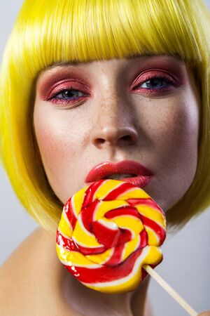 Beauty portrait of cute young girl with freckles, red makeup and yellow wig, holding colorful candy stick on lips and looking at camera with passion . indoor studio shot, isolated on gray background.の写真素材