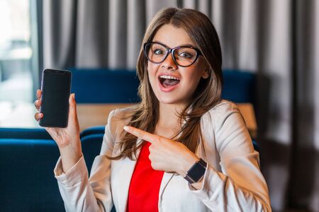 Portrait of shocked beautiful stylish young woman in glasses sitting, showing and pointing at mobile smart phone screen and looking at camera with surprised face. indoor studio shot, office backgroundの写真素材