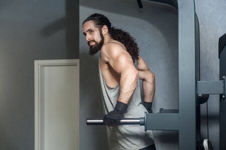 Portrait of powerful confident young adult strong man with long curly hair working out in gym,...