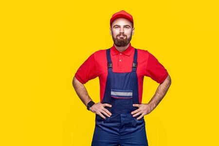 Portrait of young confident repairman with beard in blue overall, red t-shirt and cap standing and holding hands on waist with smile, indoor, studio shot, isolated on yellow background, copy space.の写真素材