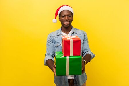 Portrait of kind handsome young man in santa hat and casual denim shirt standing looking at camera with toothy smile and giving christmas presents. indoor studio shot isolated on yellow backgroundの写真素材