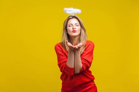 Portrait of angelic beautiful young woman with bright makeup in red casual clothes standing with closed eyes, halo on head, sending heartful air kiss. indoor studio shot isolated on yellow backgroundの写真素材