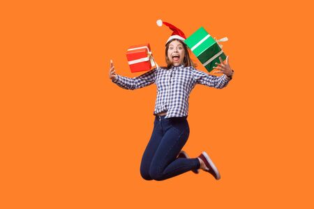 Portrait of extremely happy brunette woman in santa hat and checkered shirt jumping for joy, flying with wrapped xmas gift boxes, celebrating sales. indoor studio shot isolated on orange backgroundの写真素材