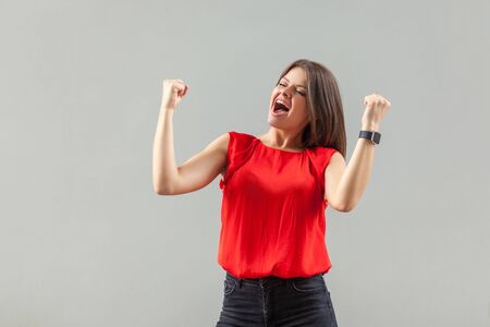 Yes! Portrait of happy beautiful brunette young woman in red shirt standing and celebrating her victory with happiness and rejoicing. indoor, studio shot, isolated on gray background.の写真素材