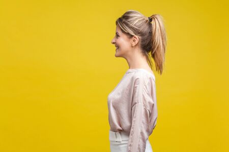 Profile side view of cheerful satisfied young woman with ponytale hairstyle and in casual beige blouse standing looking with sincere smile, positivity. indoor studio shot isolated on yellow backgroundの写真素材