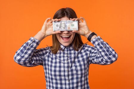 Portrait of amazed rich brunette woman wearing checkered shirt standing closing eye with dollar banknotes and smiling, surprised facial expression. indoor studio shot isolated on orange backgroundの写真素材