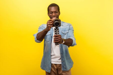 Portrait of happy photographer, joyous man in denim casual shirt holding professional digital dslr camera and taking photo, smiling enjoying his job. indoor studio shot isolated on yellow backgroundの写真素材