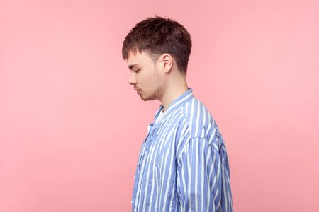 Side view portrait of upset brown-haired man with small beard and mustache in casual striped shirt standing looking down with bowed head, depression. indoor studio shot isolated on pink backgroundの写真素材