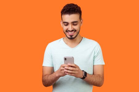 Portrait of cheerful brunette man with beard in white t-shirt using cellphone and smiling, reading good news message, enjoying mobile application. indoor studio shot isolated on orange backgroundの写真素材
