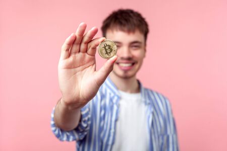 Portrait of positive excited brown-haired man in casual shirt standing showing golden bitcoin and looking at camera with toothy smile, cryptocurrency. indoor studio shot isolated on pink backgroundの写真素材