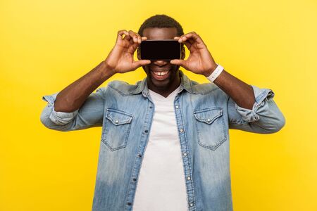 Portrait of positive cheerful man with toothy smile in denim casual shirt covering eyes with...