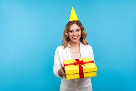 Portrait of attractive positive woman with birthday party cone on head and in white jacket holding present box and smiling at camera, greeting on holiday. studio shot isolated on blue backgroundの写真素材