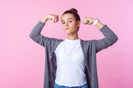 Portrait of self-confident teenage girl with bun hairstyle in casual clothes raising hands showing...