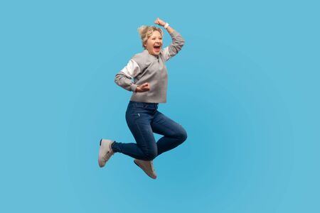 Portrait of enthusiastic happy woman with short curly hair in sweatshirt jumping in air showing yes i did it gesture, life energy and freedom concept. indoor studio shot isolated on blue backgroundの写真素材