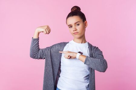 Look at my strength! Portrait of confident brunette teenage girl with bun hairstyle in casual clothes pointing at her biceps, feeling powerful and independent. studio shot isolated on pink backgroundの写真素材