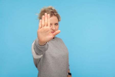 Denial, rejection concept. Young woman with short hair in sweatshirt showing stop gesture, no sign with raised palm, warning about prohibition, limit. indoor studio shot isolated on blue backgroundの写真素材