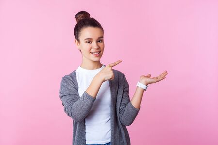 Portrait of adorable happy brunette teenage girl with bun hairstyle in casual clothes pointing to the side, holding palm up and smiling, copy space for ad. indoor studio shot isolated, pink backgroundの写真素材