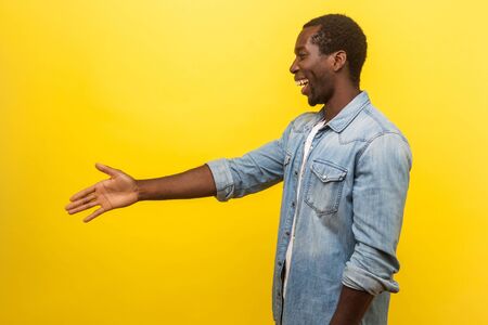 Side view portrait of friendly cheerful man in denim casual shirt with rolled up sleeves giving hand to handshake, getting to know new people, meeting. studio shot isolated on yellow backgroundの写真素材
