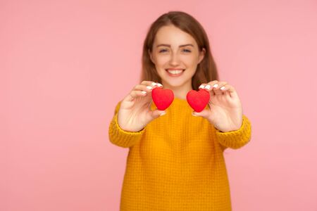 Portrait of beautiful cheerful ginger girl with charming toothy smile and in casual sweater holding little toy hearts, showing symbol of love to camera. indoor studio shot isolated on pink backgroundの写真素材