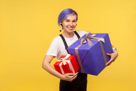 Portrait of admired joyful hipster girl with violet short hair in overalls holding two gifts, big...