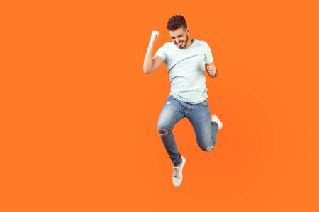 Full length portrait of ecstatic brunette man with beard in sneakers and denim outfit jumping in air showing yes i did it gesture, copy space for ad. indoor studio shot isolated on orange backgroundの写真素材