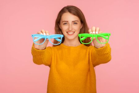 Optics and eyesight. Portrait of cheerful ginger girl in sweater holding green and blue eyeglasses and smiling at camera, recommending spectacles. indoor studio shot isolated on pink backgroundの写真素材