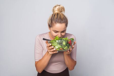 Portrait of young woman with hair bun sniffing bawl of green vegetable salad, enjoying smell of fresh healthy food, vegetarian diet, vegan nutrition. indoor studio shot isolated on grey backgroundの写真素材