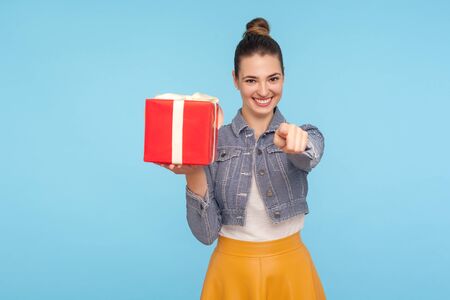 Bonus for you, winner! Portrait of optimistic beautiful fashionably dressed girl with hair bun holding gift box and pointing to camera, gesturing hey you. studio shot isolated on blue backgroundの写真素材