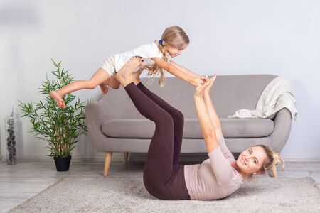 Cheerful young mother practicing yoga with little daughter, raising kid having fun and looking at camera, happy family doing gymnastics exercise together, fitness training with child at home. healthの写真素材