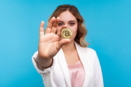 Cryptocurrency. Portrait of young woman with wavy hair in white jacket holding physical golden bitcoin, advertising of btc crypto coin, digital currency. indoor studio shot isolated on blue backgroundの写真素材