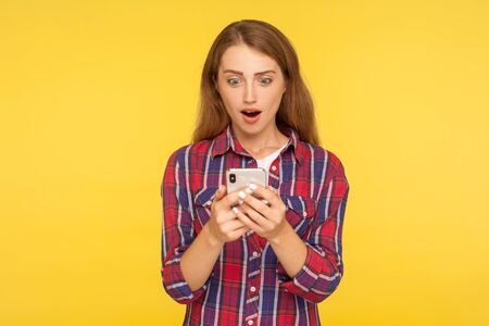 Convenient online app. Portrait of amazed ginger girl using mobile phone and looking shocked pleasantly surprised by mobile service, typing message. indoor studio shot isolated on yellow backgroundの写真素材