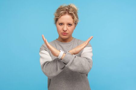 Finish, this is the end. Young woman in casual sweatshirt making x sign with crossed hands, gesturing stop prohibited, warning to ban, body language. indoor studio shot isolated on blue backgroundの写真素材