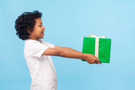 Take this present! Side view of cheerful generous good-natured little boy with curls offering gift box and smiling happily, kind child giving charity on holiday. indoor studio shot blue backgroundの写真素材