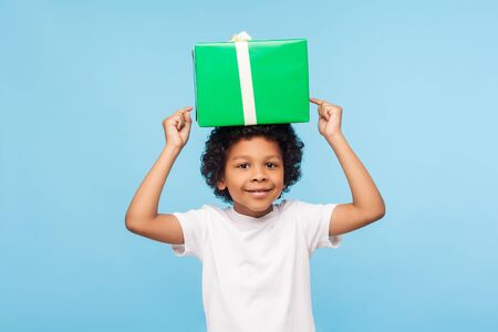 Birthday celebration. Cute joyful little boy with curly hair holding large wrapped gift box on his head and smiling happily at camera, pointing fingers at present. indoor studio shot blue backgroundの写真素材