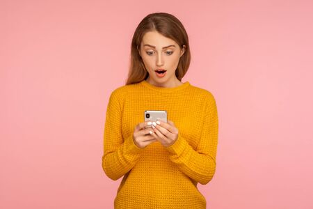 Portrait of surprised cute ginger girl texting on cellphone and expressing amazement, reading shocking message using smartphone, mobile application. indoor studio shot isolated on pink backgroundの写真素材