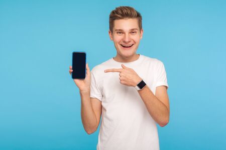 Portrait of happy joyful man in white t-shirt pointing to cellphone and looking at camera with...