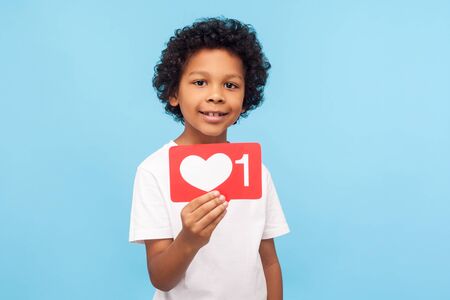 Wonderful charming little boy with curly hair holding one heart Like icon, button of social network to appreciate content, follower notification. indoor studio shot isolated on blue backgroundの写真素材