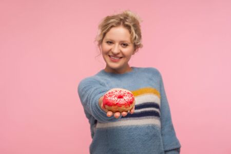 Portrait of happy adult woman with short hair in sweater giving doughnut to camera and smiling, offering tasty confection, dessert. indoor studio shot isolated on pink backgroundの写真素材