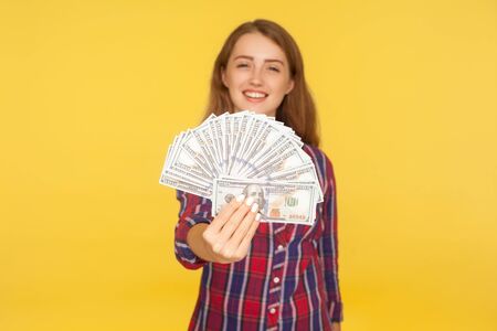 Portrait of young millionaire, rich happy girl in checkered shirt holding dollar banknotes and smiling, showing money to camera, lottery winner. indoor studio shot isolated on yellow backgroundの写真素材